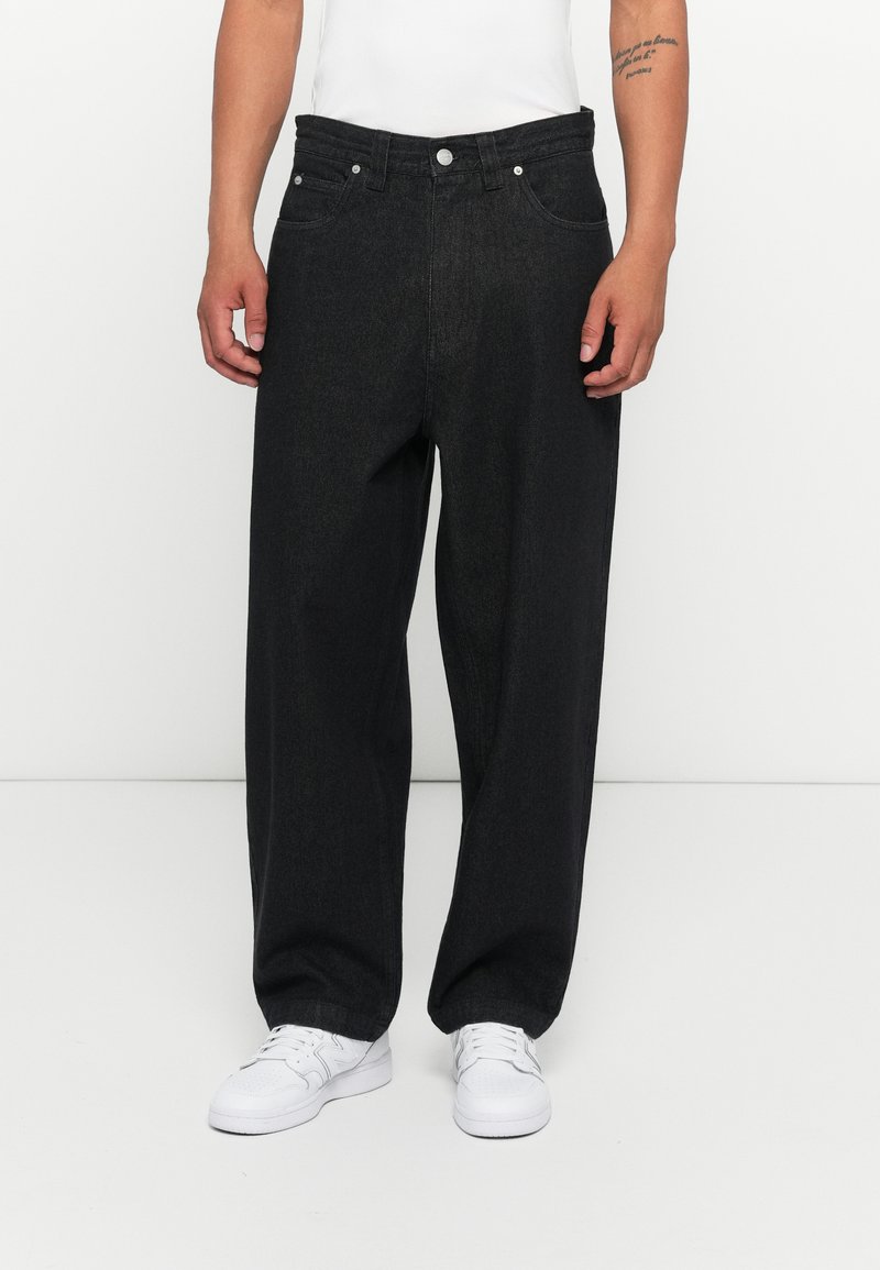 Jeans largas em denim preto com uma cintura alta, bolsos frontais e metal. Combinados com ténis brancos e uma t-shirt branca ajustada.