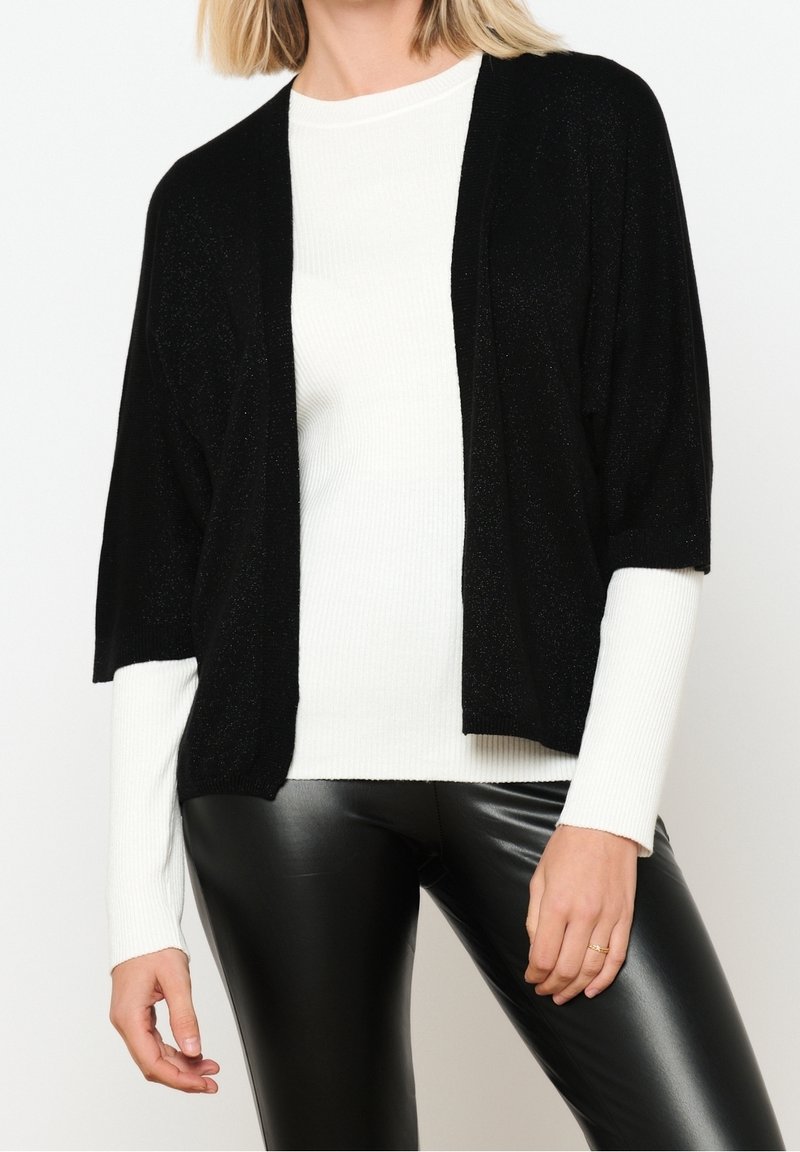 Svart, kortärmad cardigan i ett lätt, skimrande material, öppen frontdesign, lager över en vit långärmad topp.