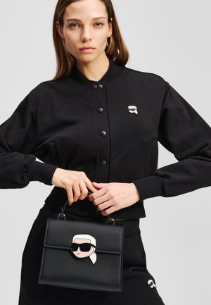 Čierna kožená kabelka s dizajnom karikatúry v bielej a čiernej farbe, s pevným tvarom a hornými uchami. Modelka nosí zodpovedajúci outfit.