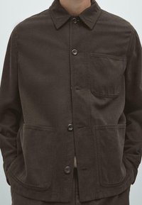 Donkerbruine jack met een kraag, knopen aan de voorkant, twee grote voorzakken en een gestructureerde stof. Minimalistisch ontwerp zonder zichtbare patronen.