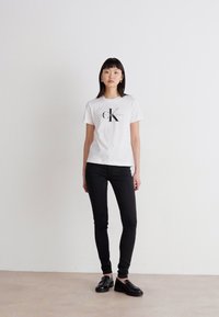 Witte katoenen t-shirt met zwarte grafische "CK Jeans" logo, gecombineerd met zwarte skinny jeans en zwarte schoenen, op een model tegen een neutrale achtergrond.
