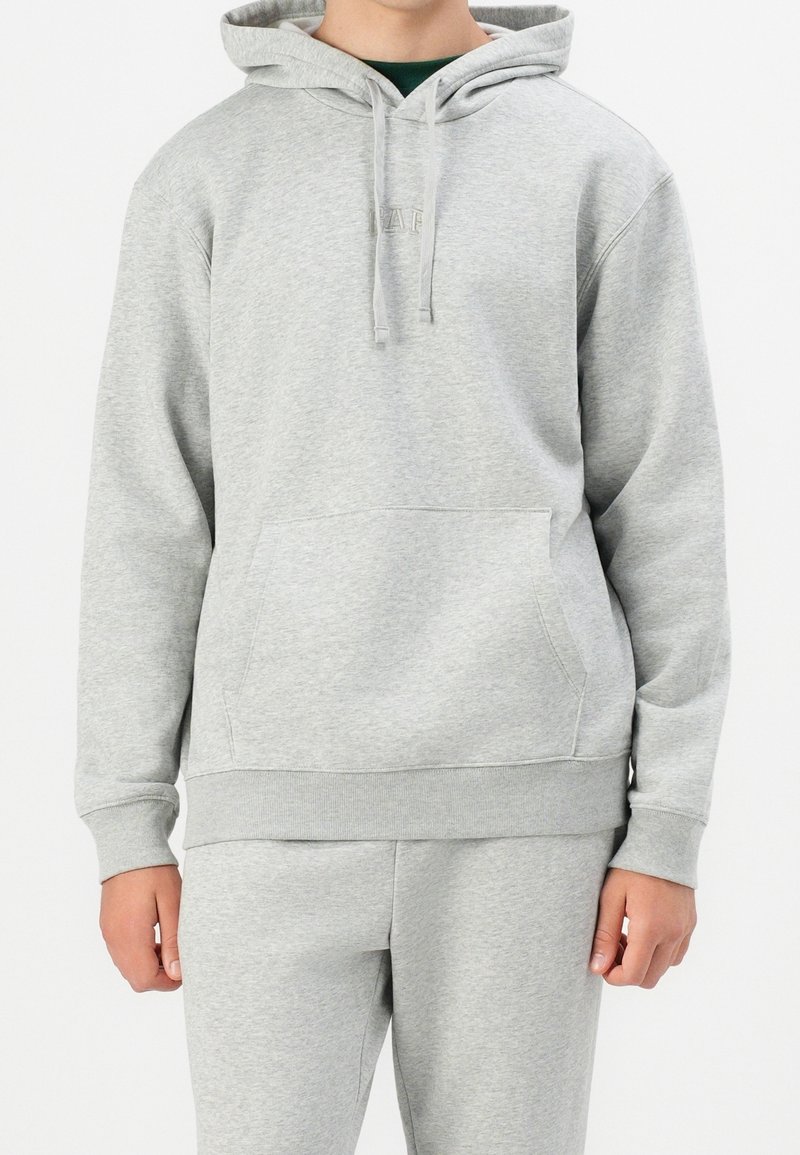 Homme portant un sweat à capuche gris clair et un pantalon de survêtement assorti, les mains détendues le long du corps sur fond blanc.