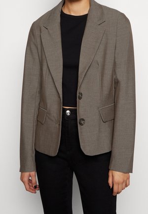 Personne portant un blazer taupe cintré à deux boutons, sur un top court noir et un pantalon noir taille haute, debout devant un fond uni.