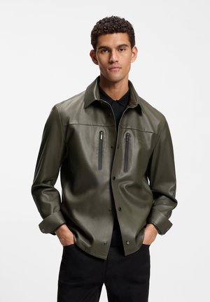 Ung mand iført en mørkegrøn læderjakke med to lodrette brystlommer med lynlås, sort skjorte og sorte bukser, hænderne i lommerne.