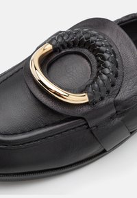 Gros plan d'un mocassin en cuir noir avec une grande boucle circulaire en cuir tressé et doré sur le dessus de la chaussure.