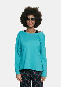 Turkusowy oversize'owy sweter z opadającymi ramionami. Czarne spodnie z kolorowym geometrycznym wzorem. Modelka nosi ciemne okulary przeciwsłoneczne.