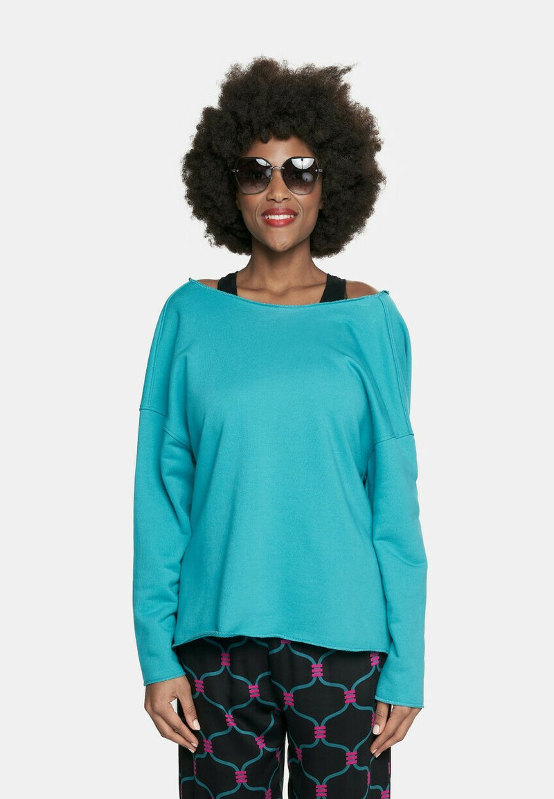 Turkusowy oversize'owy sweter z opadającymi ramionami. Czarne spodnie z kolorowym geometrycznym wzorem. Modelka nosi ciemne okulary przeciwsłoneczne.