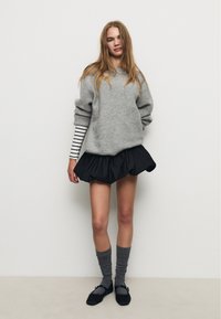 Jeune femme portant un pull gris oversized, une chemise rayée à manches longues en dessous, une jupe noire, des chaussettes grises et des chaussures Mary Jane noires, se tenant contre un mur blanc.