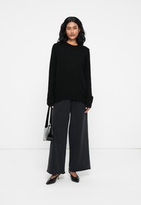 Suéter oversized preto, calças largas escuras, sapatos pretos de bico fino e uma pequena bolsa preta. Textura suave, ajuste descontraído.