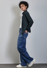 Giacca zip-up blu scuro sopra una maglietta bianca, abbinata a jeans chiari a vestibilità ampia. Modello indossa sneakers bianche. Sfondo semplice.