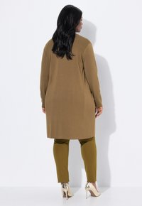 Frau mit langen dunklen Haaren, die einen braunen knielangen Mantel, olivgrüne Hosen und goldene High Heels trägt, steht vor einer weißen Wand.