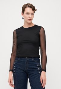 Svart langermet topp med gjennomsiktige mesh-ermer, tettsittende design, rund hals, paret med høytlivde denimjeans med snøring.