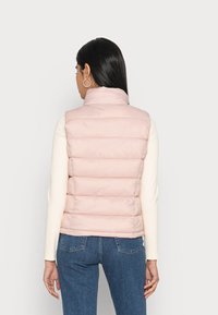Chaleco acolchado de color rosa claro con cuello alto, diseño de costuras horizontales y textura suave, acompañado de una blusa de manga larga color crema y jeans de mezclilla azul.