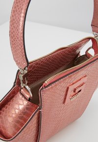 Ruusukultainen käsilaukku, jossa on teksturoitu, koholla oleva viimeistely. Varustettu vetoketj closure, brändätyt metalliosat ja yksi kantokahva.