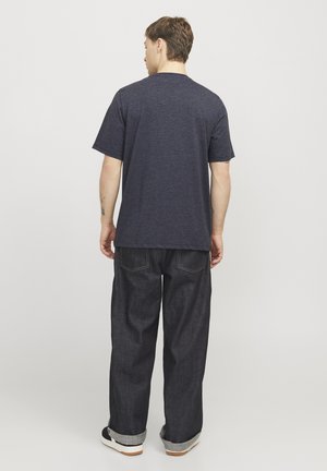 Man staat met de rug naar de kijker, draagt een donkergrijs T-shirt, losse donkere spijkerbroek met omgeslagen pijpen, en zwarte sneakers met witte zolen.