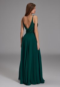 Femme portant une robe longue vert foncé avec un dos nu et des bretelles fines, debout devant un fond gris uni.