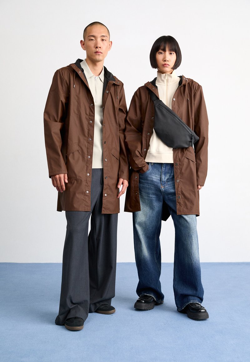 Two individuals wear brown, knee-length raincoats with hoods and button closures. One carries a black crossbody bag; both wear casual footwear.