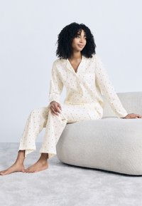 Γυναίκα με σγουρά μαλλιά κάθεται ξυπόλητη σε φωτεινό, υφασμάτινο πουφ, φοράει κρεμ πιτζάμες με μικρά κόκκινα μοτίβα καρδιών.