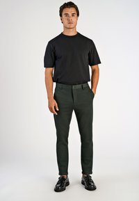 Čierna košeľa s krátkym rukávom v kombinácii s tmavozelenými, na mieru ušitými nohavicami s bočnými vreckami a manžetami na koncoch nohavíc, doplnená čiernymi elegantnými topánkami.