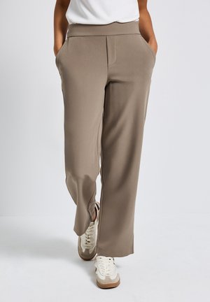 Lichtbruine wijde broek met een gladde afwerking, twee zijzakken en een hoge elastische taille. Gecombineerd met witte en beige sneakers.