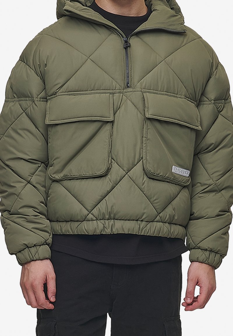 Homme portant une doudoune matelassée vert olive avec deux grandes poches à rabat à l'avant et un col demi-fermeture éclair, associée à un pantalon noir.
