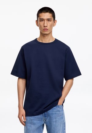 Jeune homme aux cheveux courts et foncés portant un t-shirt uni bleu marine surdimensionné et un jean bleu clair, debout sur un fond blanc.