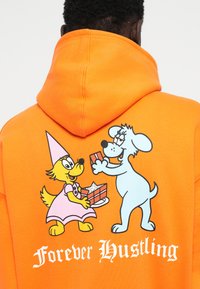 Felpa arancione con animali dei cartoni: un cane giallo in un vestito rosa che tiene un regalo e un cane blu con gli occhiali. Il testo dice "Sempre impegnati".