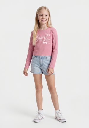 Lächelndes Mädchen mit langen blonden Haaren, das ein pinkes langärmeliges Shirt, hellblaue Jeansshorts und weiße High-Top-Sneaker trägt und steht.