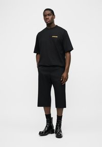 Άνδρας φοράει μαύρο oversize T-shirt με κίτρινο κείμενο, μαύρα σορτς μέχρι το γόνατο, μαύρες κάλτσες και γυαλιστερά μαύρα μποτάκια αστραγάλου, στέκεται σε λευκό φόντο.