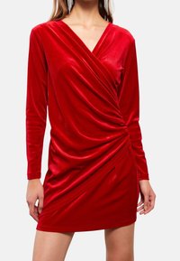 Robe en velours rouge avec des manches longues, décolleté en V profond et détails drapés à l'avant. Texture douce, silhouette ajustée et ourlet à hauteur des genoux.