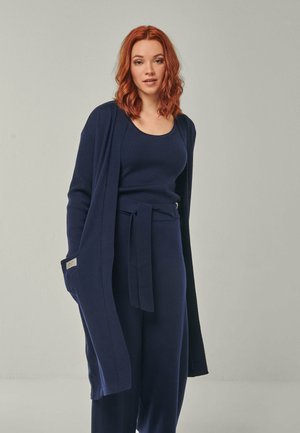 Frau mit schulterlangen roten Haaren, trägt ein marineblaues Tanktop, eine hose mit Gürtel und eine lange Strickjacke vor hellem Hintergrund.