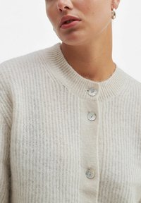 Beige ribbestrikket cardigan med rund hals, med fire lyse knapper foran og et mykt, teksturert ullblandingsstoff.