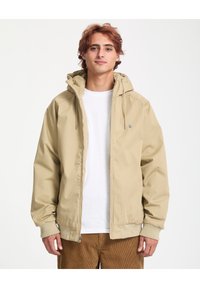 Giacca beige con cappuccio, zip completa, polsini elastici e coulisse. Indossata sopra una maglietta bianca, abbinata a pantaloni di velluto a coste.