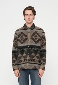 Chaqueta camisa de lana con un patrón geométrico en tonos marrón, negro y gris, cierre frontal con botones y cuello negro.