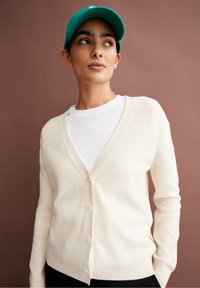 Cremfarbener Strickcardigan mit V-Ausschnitt, fünf Knöpfen und ribbed Bündchen. Über einem weißen T-Shirt getragen. Verfügt über eine glatte Textur und ein lässiges Design.
