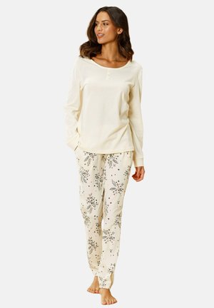 Frau trägt cremefarbenes langärmeliges Oberteil und cremefarbene, mit Blumen gemusterte Pyjamahose, steht barfuß vor einfarbigem Hintergrund.