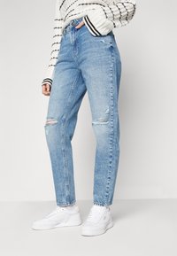 Blå denimjeans med hög midja, som har slitna partier och en avslappnad passform, parat med vita sneakers. Den randiga tröjan ger kontrast.