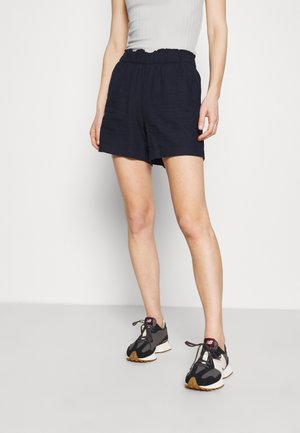 Bermudas de lino azul marino con cintura elástica. Combinadas con zapatillas de correr en negro y crema con suela texturizada y diseño de cordones.