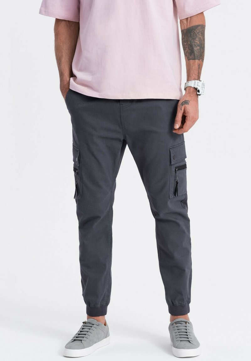 Cargo broek in donkergrijze stof met twee zijzakken en elastische boorden, gecombineerd met een lichtroze shirt en grijze sneakers.