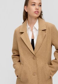 Jonge vrouw in een camelkleurige jas over een witte shirt en zwarte top, met haar handen in de zakken, die iets opzij kijkt tegen een effen achtergrond.