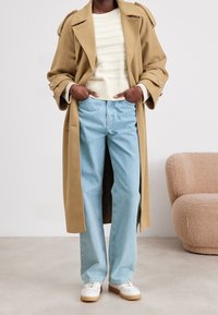 Abrigo trench beige con grandes solapas, suéter de canalé crema, jeans rectos azul claro y zapatillas blancas con suela crema. Fondo texturizado.