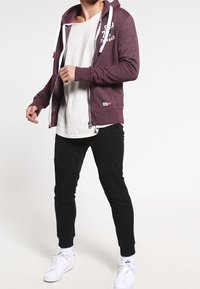 Sudadera con cremallera color burdeos con cordones blancos y logo en el pecho, combinada con una camiseta gris claro y pantalones de chándal negros ajustados, terminada con zapatillas blancas.