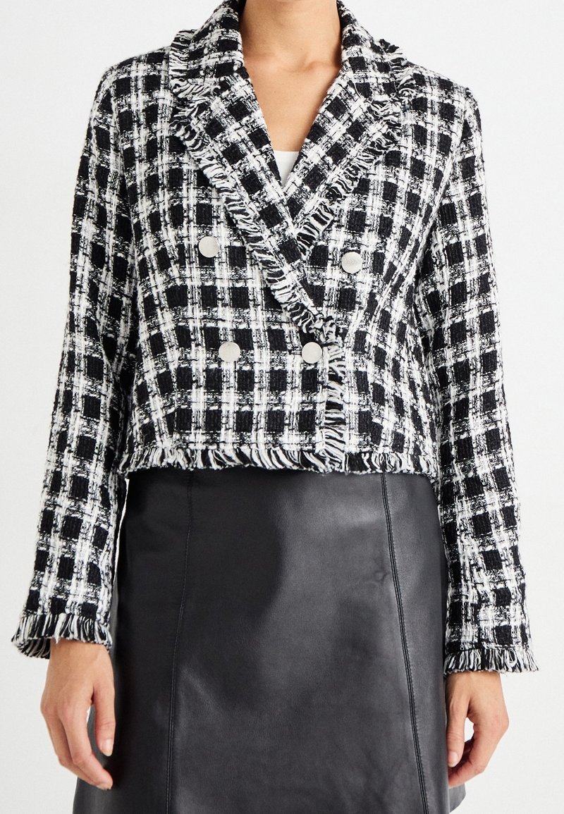 Femme portant une veste à carreaux noire et blanche texturée à double boutonnage avec des détails frangés et une jupe en cuir noire.