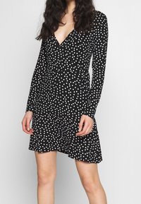 Svart omlottklänning med långa ärmar, prydd med vita prickar. Mjukt tyg med viss stretch, v-ringning och figurnära midja.
