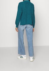 Azurově modrý pletený svetr s žebrovanou strukturou, v kombinaci s světlými modrými kalhotami širokého střihu a bílými teniskami se zelenými pruhy.