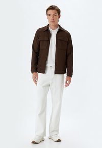 Chaqueta marrón con canalé y dos bolsillos en el pecho, llevada sobre una camiseta polo blanca y combinada con pantalones blancos. Tejido texturizado con un diseño estructurado.