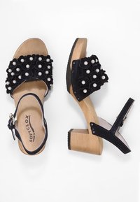 Sandali neri in suede con tacchi in legno robusti, decorazioni floreali e dettagli in perle. Cinturino regolabile alla caviglia per una calzata sicura.