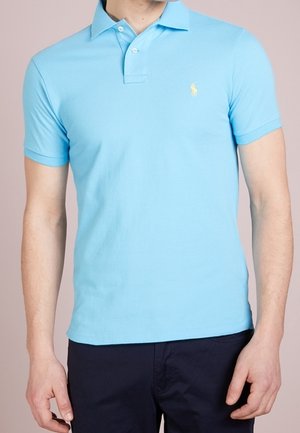 Jasnoniebieska koszulka polo z kołnierzykiem i krótkimi rękawami; posiada małe żółte logo na lewej piersi. Wykonana z gładkiej bawełnianej tkaniny.