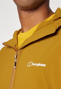 Žltohnedá bunda na zips s kapucňou a bielym logom "berghaus". Má hladkú textúru a kontrastný zips.