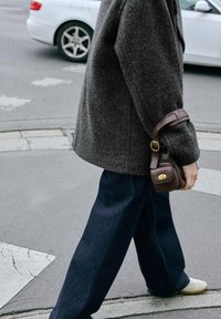 Abrigo de lana gris oscuro, jeans anchos de color navy, zapatos de tono claro. Pequeño bolso de cuero marrón con herrajes en tono dorado. Entorno urbano.